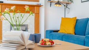 Ein Wohnzimmer mit einem blauen Sofa, einem gelben Kissen, einem Couchtisch aus Holz, einer Schale Erdbeeren und einer Vase mit Blumen.