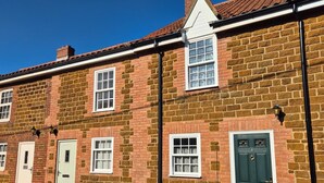 A row of brick houses with white-framed windows and doors, a clear blue sky, and a visible street. - 2 Bed in Snettisham (oc-d31668) (Snettisham)