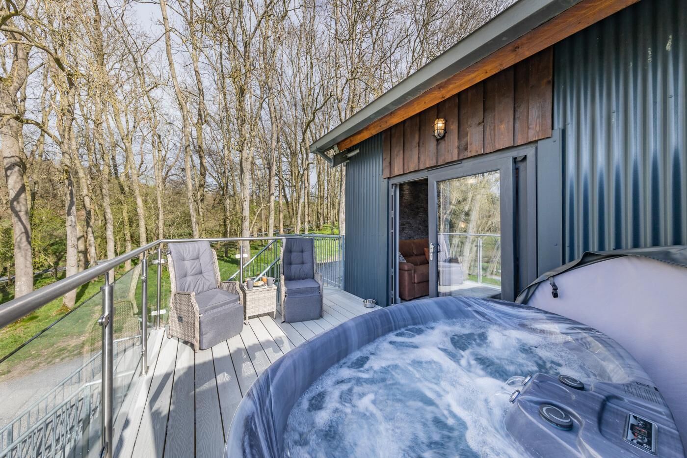 A hot tub on a deck with wicker chairs and a glass railing, overlooking a forested area.