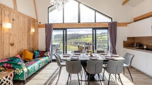 A dining area with a table set for four, a sofa with colorful cushions, and a view of the countryside through large windows. - 4 Bed in Berriew (oc-t31114) (Berriew)