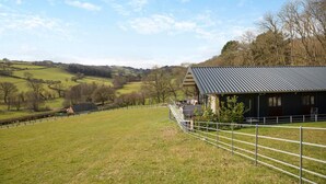 A rural property with a metal-roofed building, a fenced field, and rolling hills in the background. - 4 Bed in Berriew (oc-t31114) (Berriew)