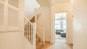 A hallway with a carpeted staircase, a white door leading to a living room with a sofa and a chandelier. - 2 Bed in Swanage (oc-b31105) (Swanage)