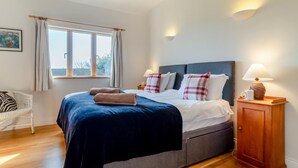 A neatly made bed with a dark blue bedspread and white linens, a wooden nightstand with a lamp, and a window with curtains. - 3 Bed in South Cove (oc-r30975) (South Cove)