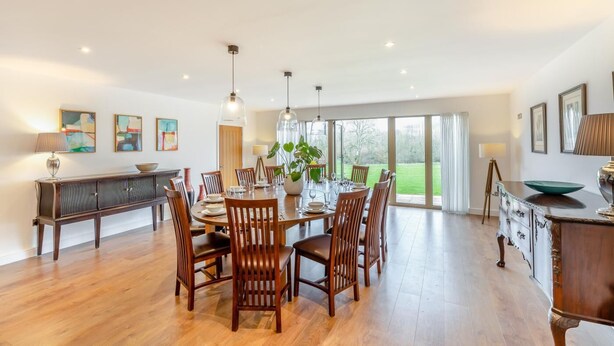 Een ruime eethoek met een grote houten tafel, meerdere stoelen en hanglampen. Er is uitzicht op een groen landschap door grote ramen.