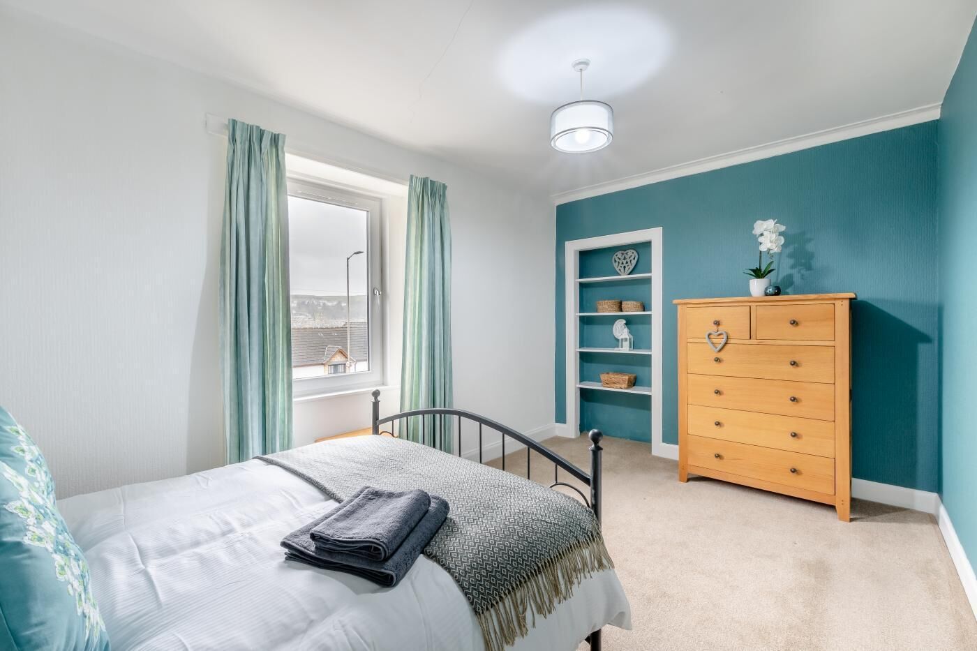 Un dormitorio con cama, cómoda, ventana con cortinas y repisa con objetos decorativos.