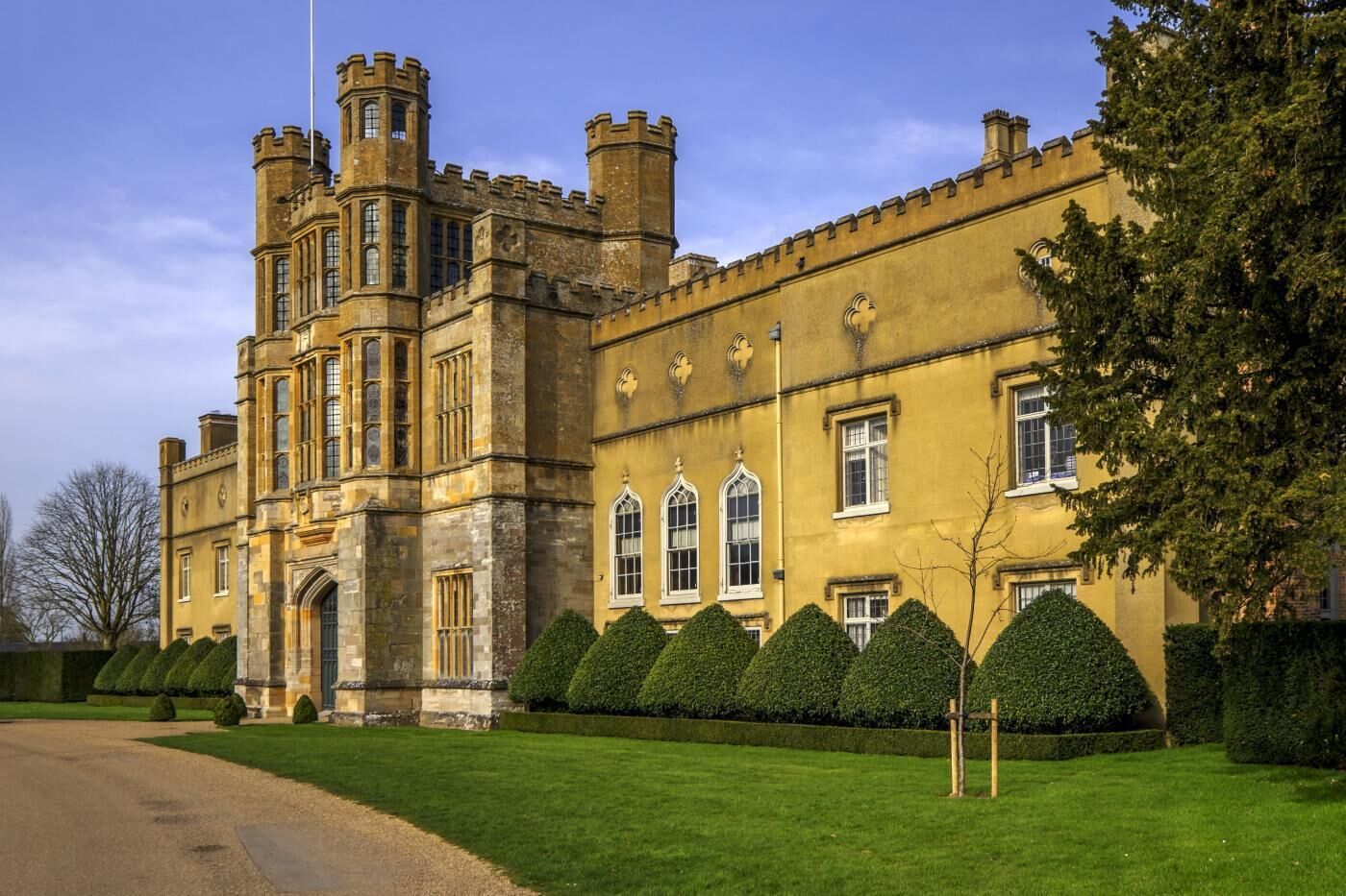 A large, historic building with a prominent tower and multiple windows, surrounded by well-maintained hedges.