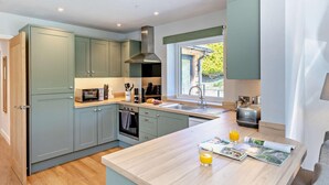 A modern kitchen with light green cabinets, a central island with a magazine and glasses, and a window providing a view of greenery outside. - 2 Bed in Llanarmon Dyffryn Ceiriog (oc-b31110) (Llanarmon Dyffryn Ceiriog)