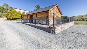 A log cabin with a stone foundation, a gravel driveway, and a fenced yard. - 2 Bed in Llanarmon Dyffryn Ceiriog (oc-b31110) (Llanarmon Dyffryn Ceiriog)