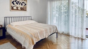Ein Schlafzimmer mit einem Bett, Holzboden, einem Fenster mit leichtem Vorhang und einem Gemälde an der Wand.