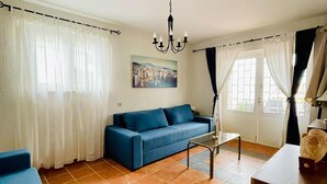 Ein Wohnzimmer mit einem blauen Sofa, einem Glastisch und einer Chandelüster. An der Wand hängt ein Gemälde und ein Fenster ist mit Vorhängen dekoriert.