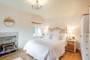 Un dormitorio con chimenea, una cama con ropa de cama blanca, una mesita de noche con lámpara y un espejo montado en la pared.