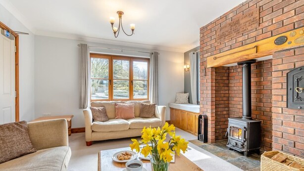 A cozy living room with a sofa, a coffee table with flowers, a fireplace, and a view through large windows. - 5 Bed in Ystrad Meurig (oc-r31133) (Ystrad Meurig)