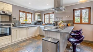 A modern kitchen with white cabinetry, a stainless steel range hood, and a central island with bar stools. - 5 Bed in Ystrad Meurig (oc-r31133) (Ystrad Meurig)