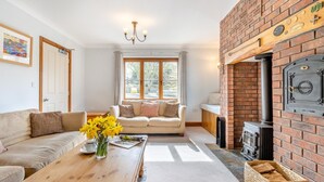 A cozy living room with a sofa, a wooden coffee table with flowers, a fireplace, and a view of the outdoors through large windows. - 5 Bed in Ystrad Meurig (oc-r31133) (Ystrad Meurig)
