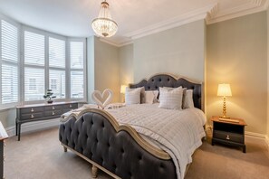 Une chambre à coucher avec un grand lit, un lustre, une table de chevet, une commode et une fenêtre avec des stores.