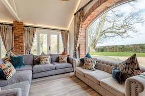 Een moderne woonkamer met een grijze zithoek, decoratieve kussens en een groot raam met uitzicht op de omgeving.