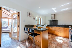 A modern kitchen with a central island, bar stools, and a dining area with a sofa and chairs. - 3 Bed in Brockenhurst (oc-w30959) (Brockenhurst)