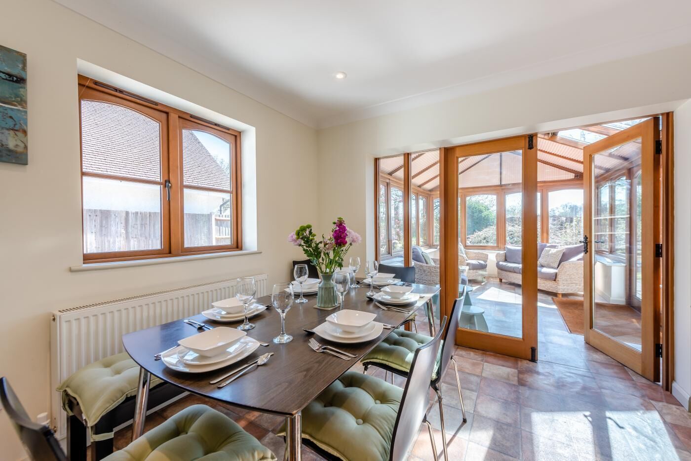 A dining area with a long table set for six, a vase with flowers, and a view through glass doors to an outdoor seating area.