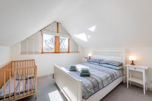 Ein Schlafzimmer mit schrägverdeckter Decke, einem Babybett, einem Bett mit gemusterten Bettwaren, zwei Nachttischen mit Lampen und einem Fenster mit Jalousien.