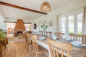 A dining area with a brick fireplace, wooden chairs, a long table set for six, and large windows with curtains. - 5 Bed in Postwick (oc-w30947) (Postwick)