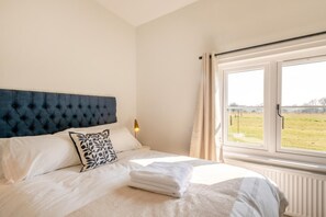 Ein ordentlich bezogenes Bett mit einer gesteppten Kopfteil, einem gemusterten Kopfkissen und Blick auf ein Feld durch ein Fenster.