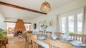 A dining area with a brick fireplace, wooden chairs, a long table set for six, and large windows with curtains. - 5 Bed in Postwick (oc-w30947) (Postwick)