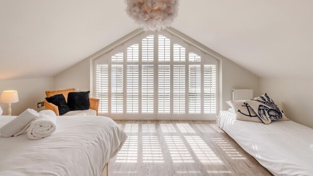 Quarto com duas camas, uma janela grande com persianas, luminária pendente e piso de madeira.