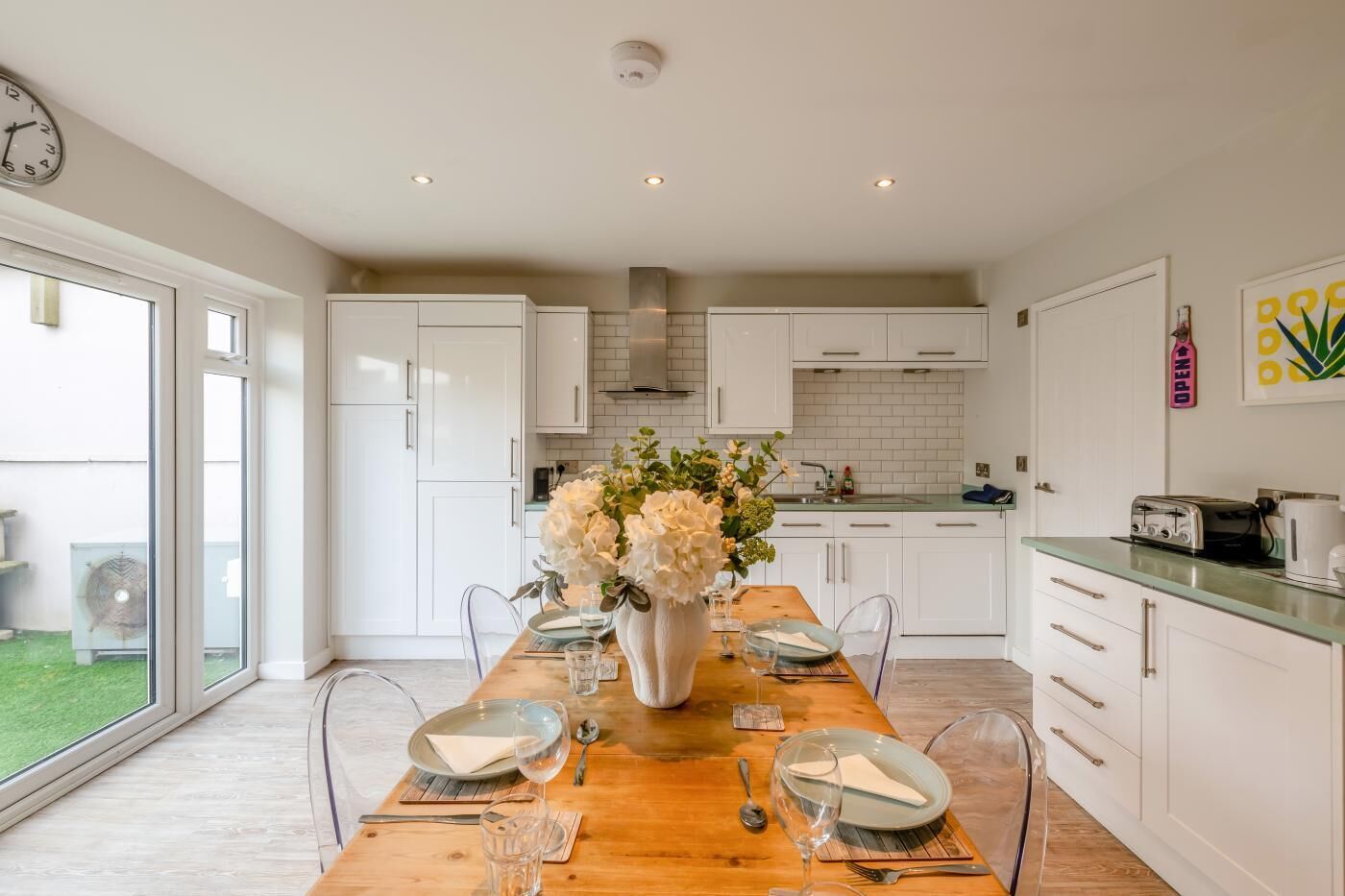 A modern kitchen with white cabinets, a wooden dining table set for four, and a large window providing a view of the outdoors.