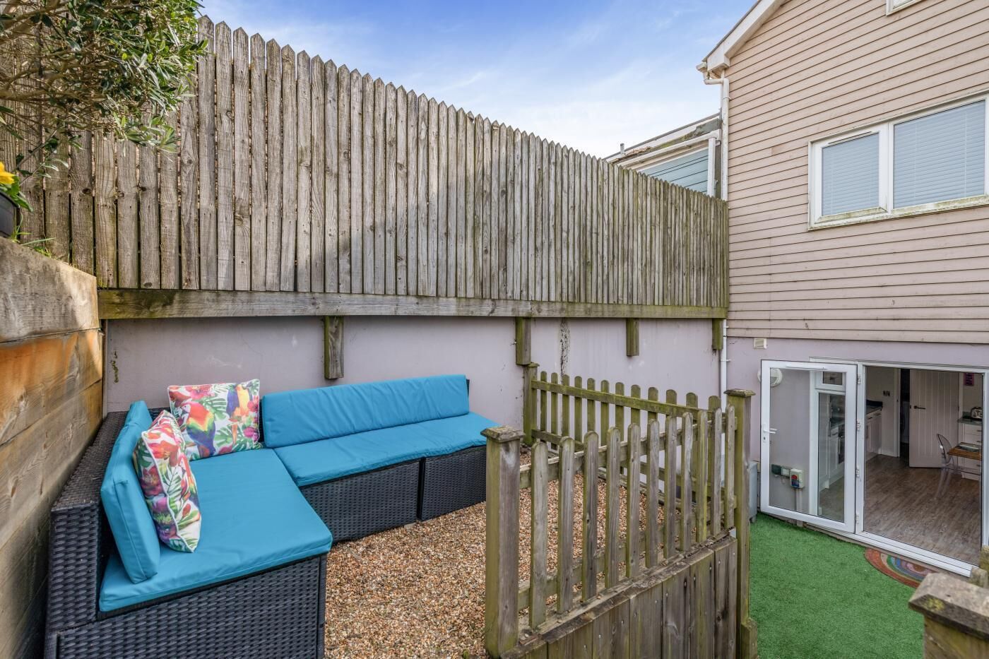 A patio area with a wicker sofa and cushions, a wooden fence, and a house in the background.