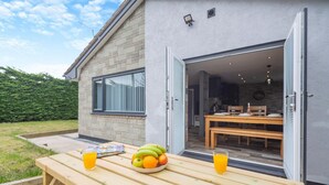 A wooden table with fruits and drinks outside a house with a brick wall and a sliding door leading to a dining area.