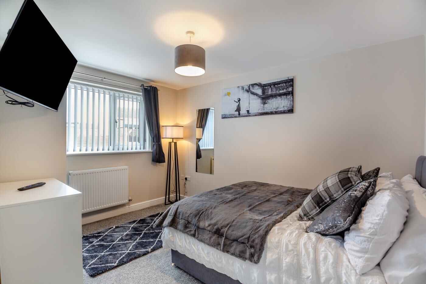 A bedroom with a bed, a desk, a standing lamp, a TV, and a window with curtains.