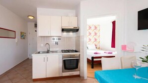 Cocina compacta con armarios blancos, estufa a gas y horno empotrado. Hay un comedor con una mesa azul y una silla, y una cama con cortina floral al fondo.