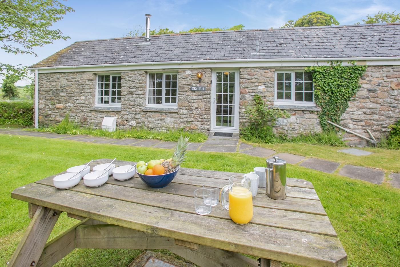 A stone building with a grassy yard, a picnic table with bowls, a fruit bowl, glasses, and a jug.