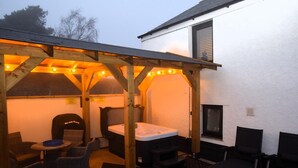 Un espace extérieur aménagé en terrasse, avec des meubles en bois, un jacuzzi et une pergola illuminée de guirlandes lumineuses.