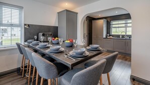 A modern dining area with a long table set for six, grey chairs, and a kitchen visible through an archway. - 3 Bed in Cockermouth (oc-o31515) (Setmurthy)
