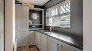 A modern kitchen with grey cabinets, a black countertop, a built-in clock, and a window with blinds. - 3 Bed in Cockermouth (oc-o31515) (Setmurthy)