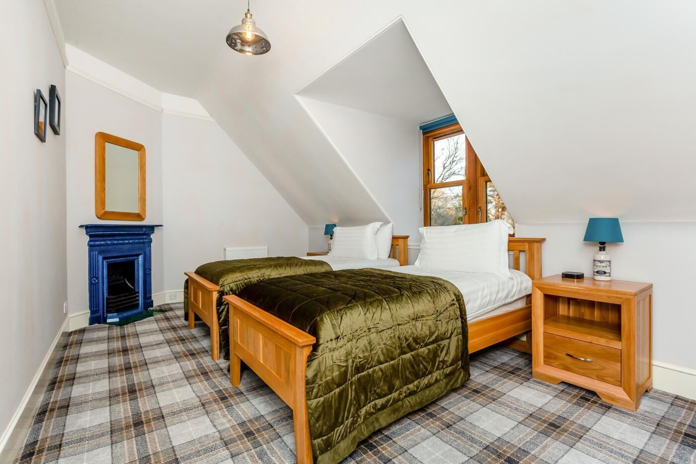 A bedroom with a sloped ceiling, a bed with a green quilt, a blue fireplace, and a nightstand with a lamp.