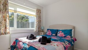 Een slaapkamer met een bed, bloemen gordijnen, een raam met uitzicht en een nachtkastje met een lamp.