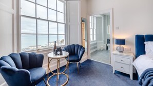 Una habitación de hotel con un ventanal grande, dos sillones, una mesita de noche, una lámpara y una cama.