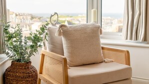A cozy room with a wicker chair, a potted plant, and a view of a cityscape through a window. - 4 Bed in St. Ives (oc-p31118) (Saint Ives)