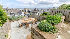 A patio with a wooden table and chairs, surrounded by a wooden fence and a garden bed. - 4 Bed in St. Ives (oc-p31118) (Saint Ives)