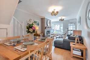 Ett matområde med ett träbord, stolar och en vas med blommor. I vardagsrummet finns en soffa och en lampa.