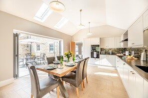A modern kitchen with white cabinets, a wooden dining table, and a view of a stone building outside. - 3 Bed in Longhorsley (oc-d31795) (Longhorsley)