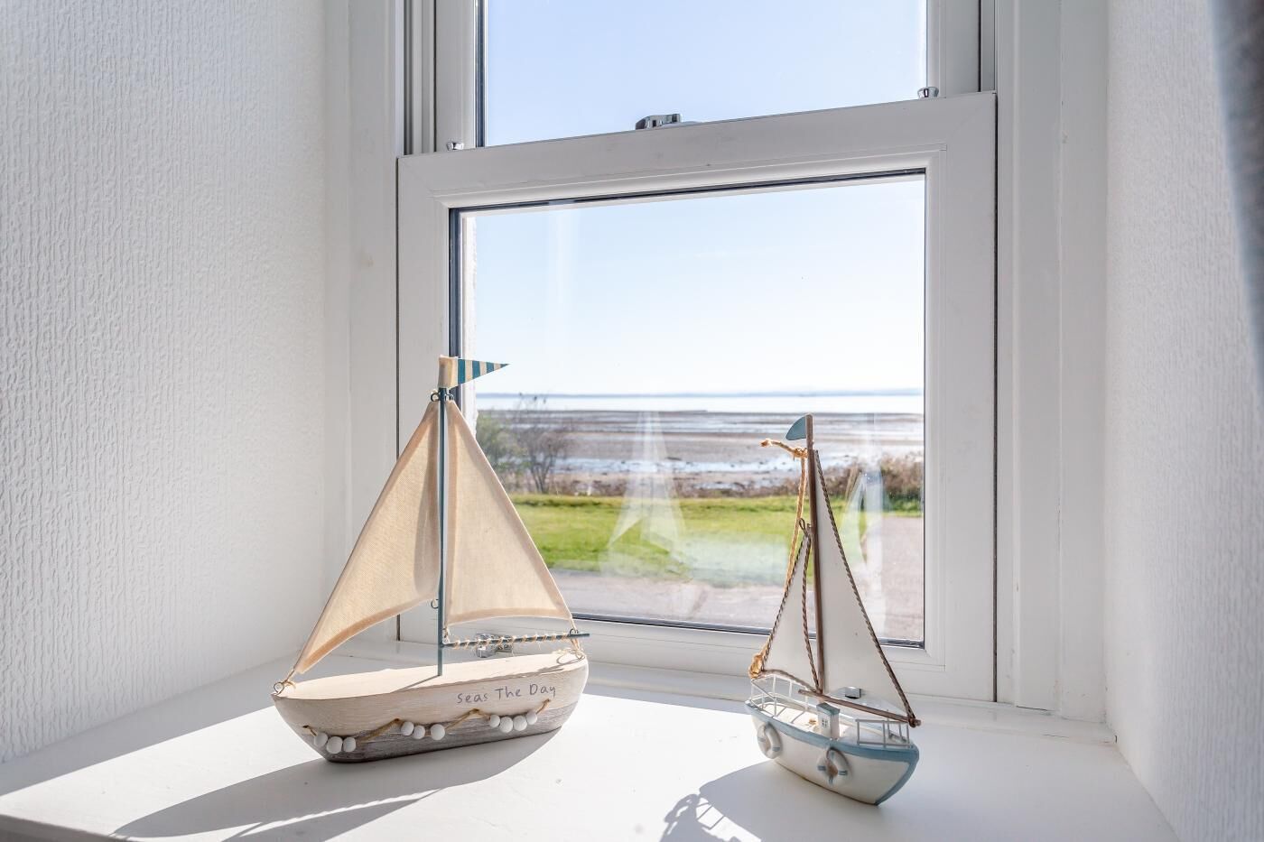 Two model sailboats on a windowsill with a view of a beach and sea.