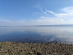 En strand med småsten, lugnt vatten och en klar himmel.