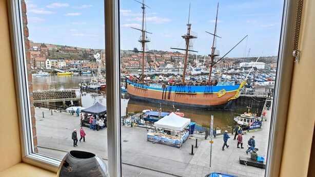 A view of a harbor with a large sailing ship docked, a market area with stalls, and a blue car parked on the street. - 2 Bed in Whitby (oc-h31801) (Whitby)