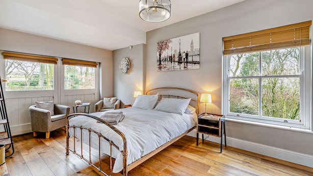 Een slaapkamer met een houten bed, twee nachtkastjes, een stoel en een groot raam met uitzicht op bomen.