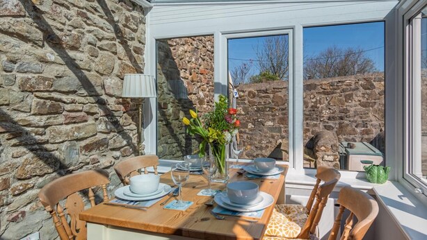 A dining area with a stone wall, wooden table, chairs, and a window with a view of a stone wall and trees. - 2 Bed in Little Haven (oc-i31766) (Little Haven)