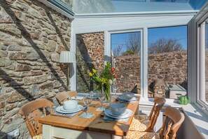 A dining area with a stone wall, wooden table, chairs, and a window with a view of a stone wall and trees. - 2 Bed in Little Haven (oc-i31766) (Little Haven)