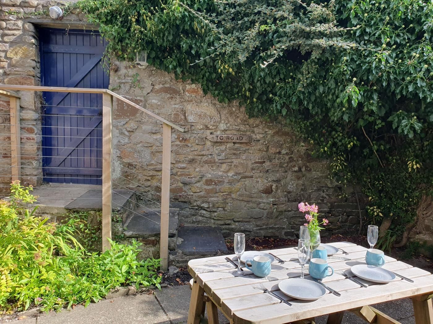 A stone wall with a wooden gate labeled "TO ROAD", a wooden table set with dinnerware and glasses, and a small garden area with plants and a pink flower vase.